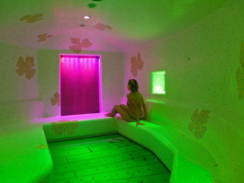 Žena sedí v světle zelené parní místnosti s růžově osvětleným oknem.