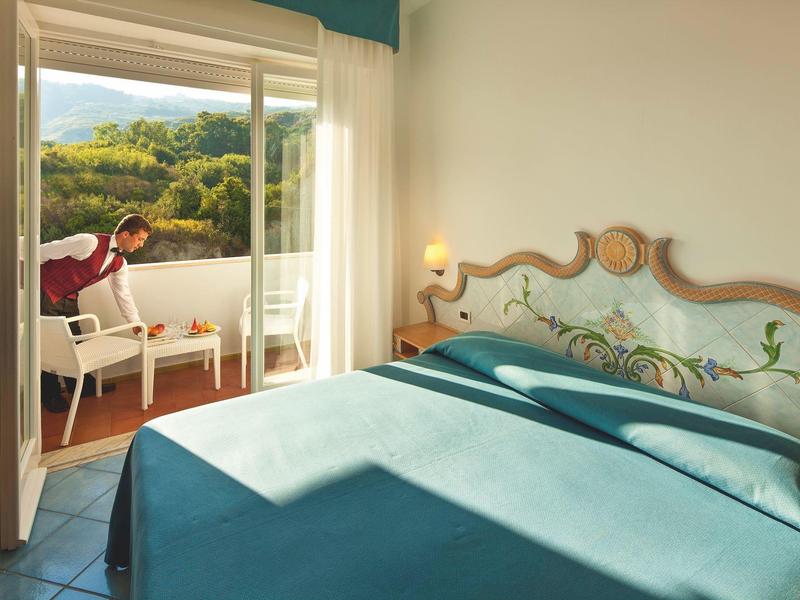 Světle zařízená ložnice s modrou přikrývkou a balkonem s výhledem na zalesněné kopce.