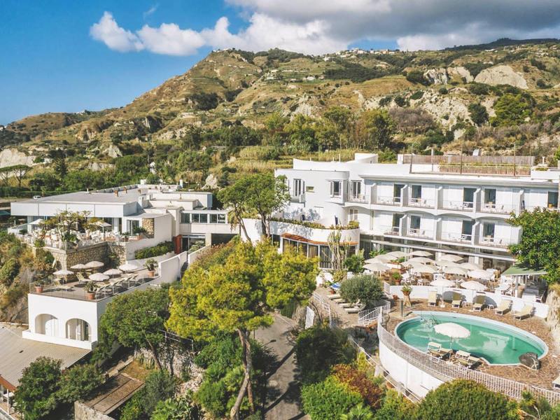 Hotelový komplex s kulatým bazénem a několika bílými budovami před zelenými horami pod modrou oblohou