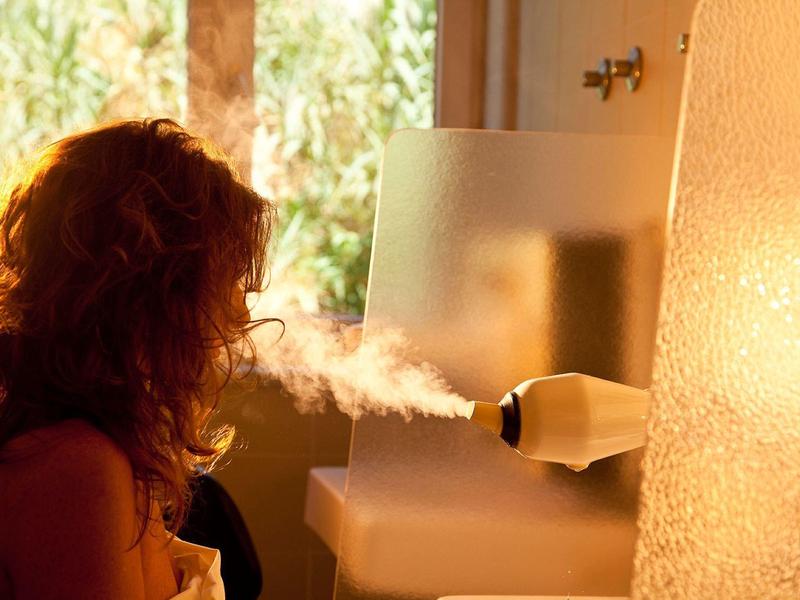 Žena používá parní zařízení na obličej v koupelně s teplým osvětlením.