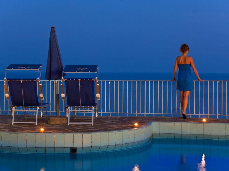 Žena stojí na okraji bazénu s výhledem na modré moře za soumraku