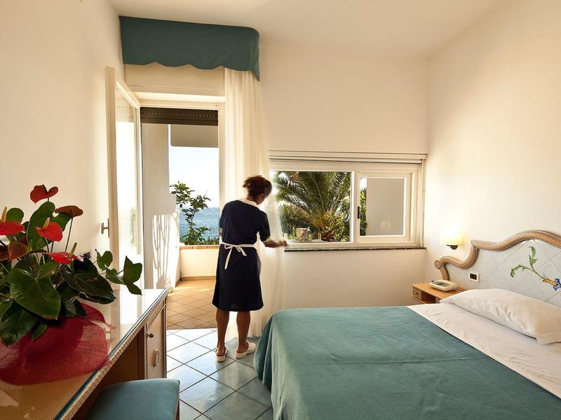 Světly hotelový pokoj s manželskou postelí a ženou u otevřeného okna s výhledem na palmy.