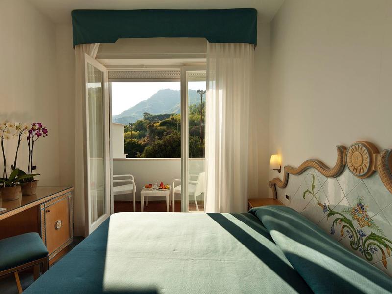 Světý hotelový pokoj s dvoulůžkem, balkonem a výhledem na hory za denního světla.