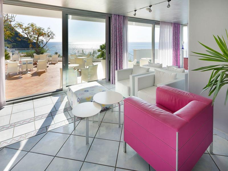 Světý pokoj s růžovou pohovkou, bílými křesly a výhledem na terasu a moře.