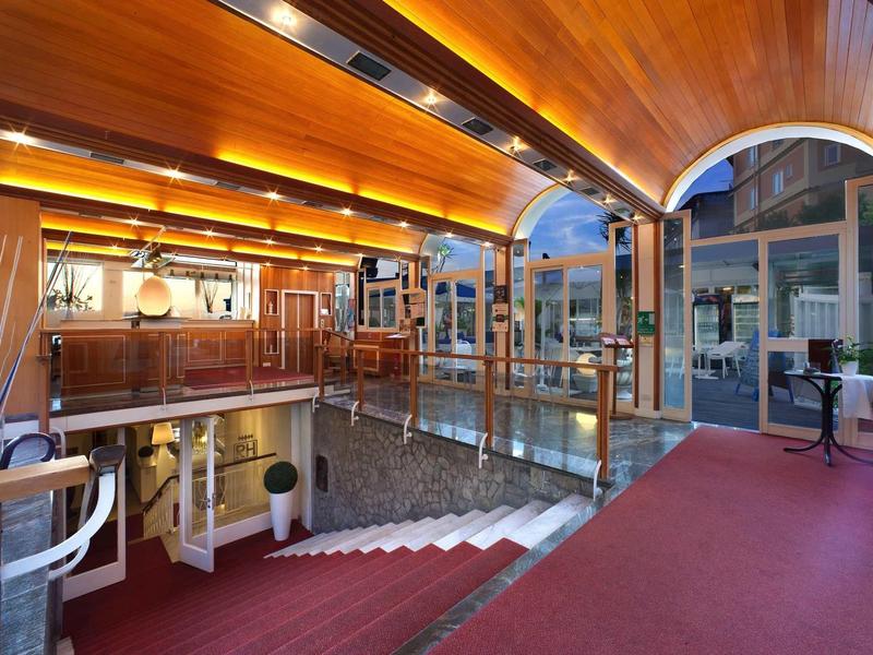 Elegancki hol hotelowy z czerwonym dywanem, szklanymi balustradami i ciepłym drewnianym wystrojem pod sklepionym sufitem.