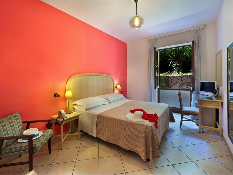 Przytulny pokój hotelowy z podwójnym łóżkiem, czerwoną ścianą akcentową i balkonem z widokiem na zieleń.