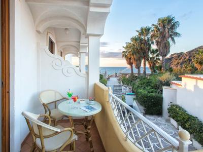 Balkon mit zwei Stühlen, Tisch mit Getränken, Blick auf Palmen und Meer im Hintergrund.