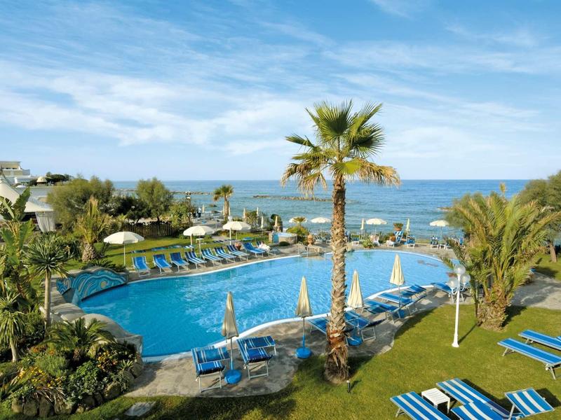 Piscina dell'hotel con lettini e palme vicino al mare sotto un cielo azzurro.
