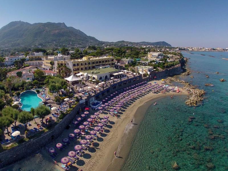 Hotel sulla spiaggia con ombrelloni rosa e acqua di mare limpida lungo la costa sotto colline boscose.