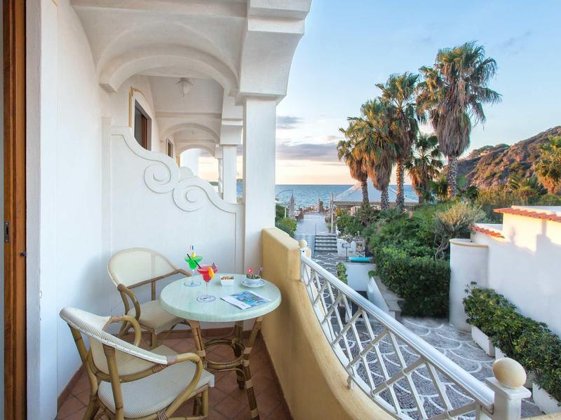 Balcone con tavolo e sedie, vista su palme e mare sullo sfondo al tramonto.
