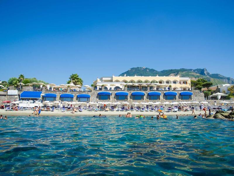 Hotel sulla spiaggia con ombrelloni blu e bianchi e acqua limpida in primo piano.