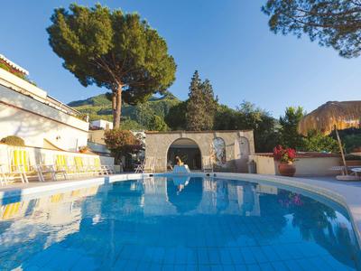 Großer blauer Pool mit weißen Liegen, umgeben von Bäumen und Gebäuden bei klarem Himmel.