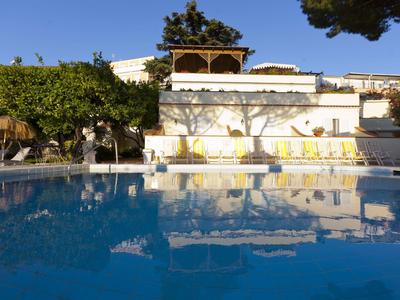 Großer blauer Pool mit weißen Liegestühlen vor weißem Gebäude und grünem Bewuchs.