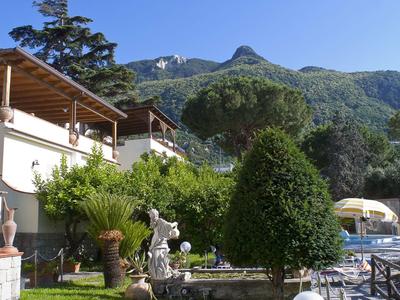 Garten mit Skulptur, Bäumen und Wohnhäusern vor bewaldeten Bergen unter blauem Himmel.