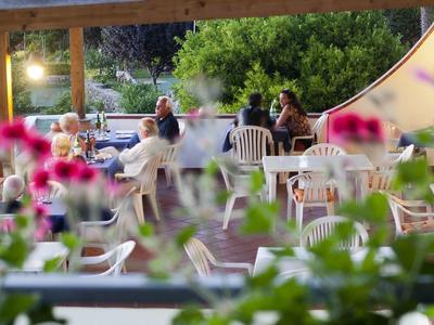 Menschen sitzen an weißen Tischen draußen, umgeben von grünen Pflanzen und pinken Blumen.