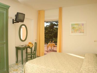 Helles Schlafzimmer mit Doppelbett, grünem Schrank, kleinem Tisch, ovalem Spiegel und gelben Vorhängen.