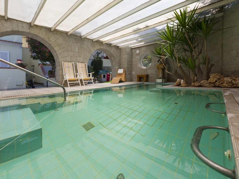 Hell beleuchtetes Hallenbad mit Pflanzen, Steinmauer und großen runden Fenstern unter Glasdach.