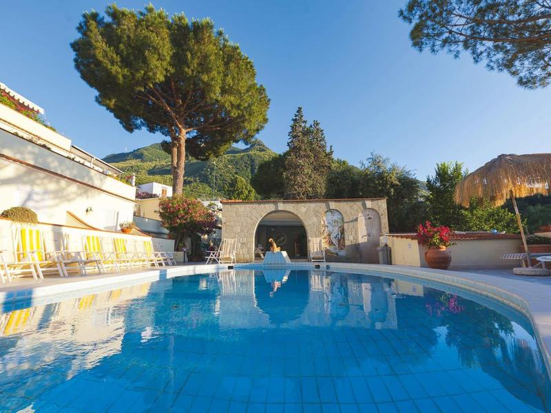 Großer blauer Pool mit weißen Liegen, umgeben von Bäumen und Gebäuden bei klarem Himmel.