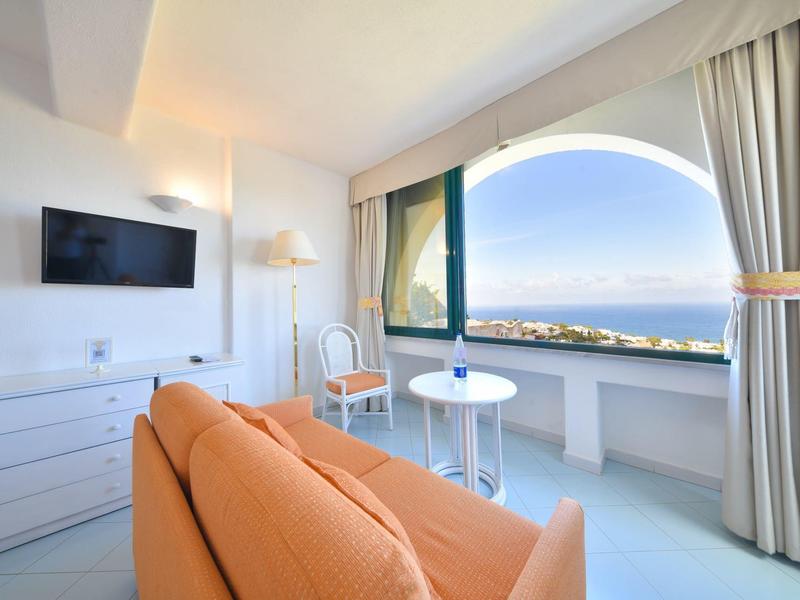 Světlý obývací pokoj s oranžovou pohovkou, bílým nábytkem a velkým oknem s výhledem na moře.