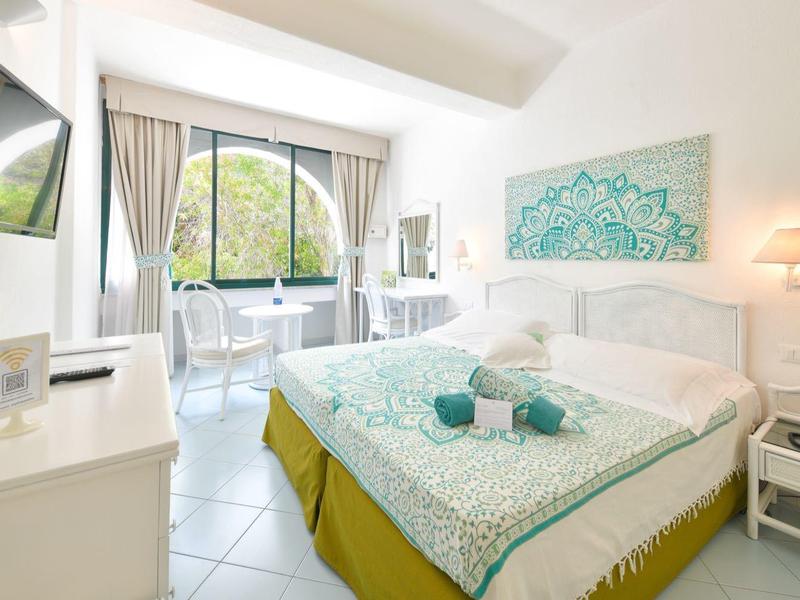 Světle hotelový pokoj s manželskou postelí, TV, psacím stolem a výhledem na zelené stromy