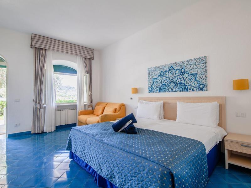 Jasný hotelový pokoj s manželskou postelí, modrým přehozem, oranžovou pohovkou a velkými okny.