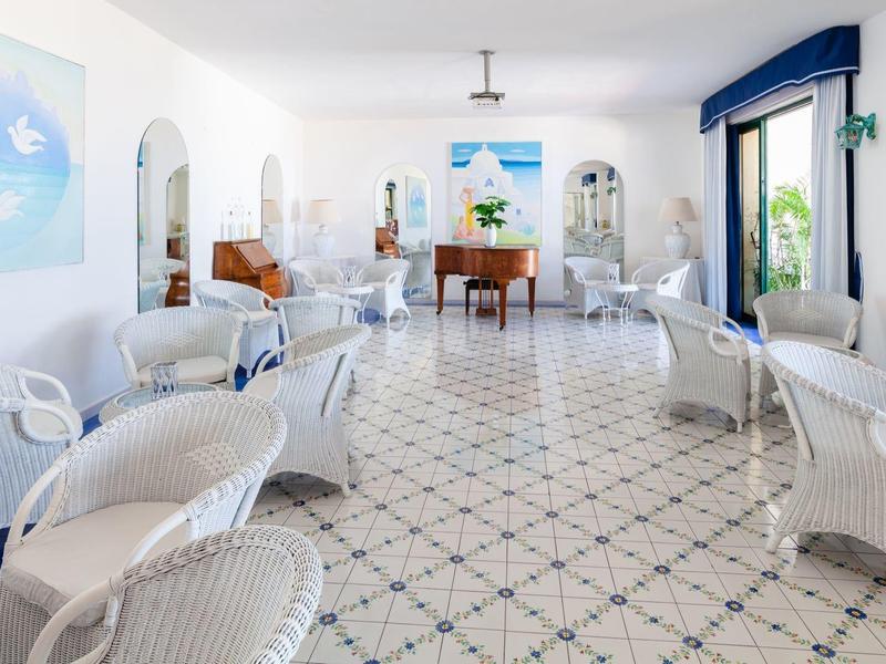 Jasná hotelová lobby s bílým ratanovým nábytkem a modrými akcenty, velké okno s výhledem ven