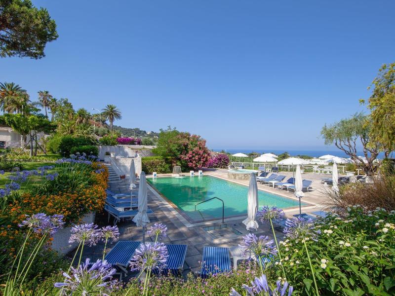 Bazén s modrými lehátky a slunečníky v středomořské zahradě s výhledem na moře.