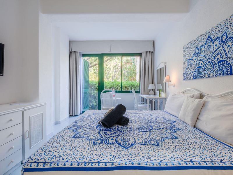 Světlo hotelový pokoj s manželskou postelí, modro-bílým vzorem, balkonem a výhledem do zeleně.