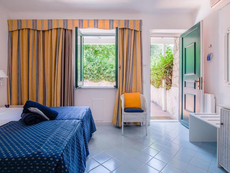 Světle osvětlený hotelový pokoj se dvěma postelemi, tyrkysovými závěsy a otevřeným oknem.