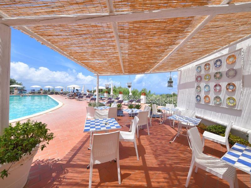 Venkovní restaurace s bílými stoly a židlemi vedle bazénu, dekorativní talíře na stěně.