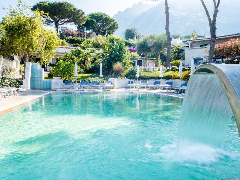 Čistá voda v bazénu s vodopádem a posezením v slunečném hotelovém prostředí se zelenými rostlinami.