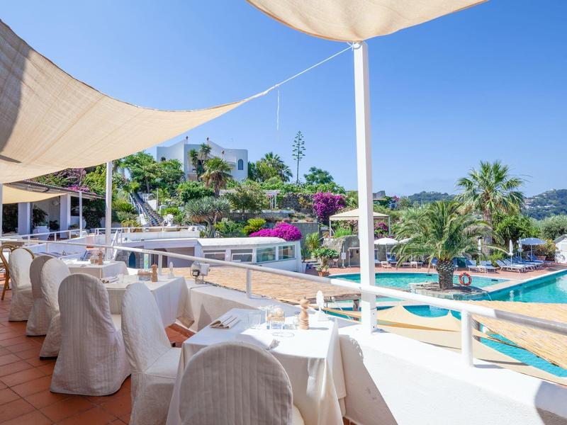 Terasa s prostřenými stoly s výhledem na bazén s palmami a jasně modrou oblohou.