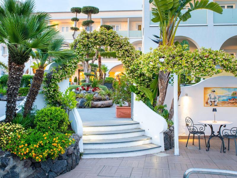 Scalinata bianca con piante verdi accanto a zona pranzo nel giardino dell’hotel alla luce della sera.