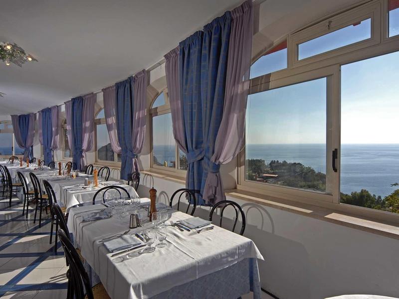 Elegant restaurant met witte tafelkleden en zeezicht door grote ramen.