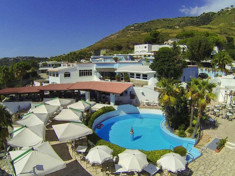 Hotelcomplex met zwembad, parasols en omliggende heuvels onder een heldere lucht.