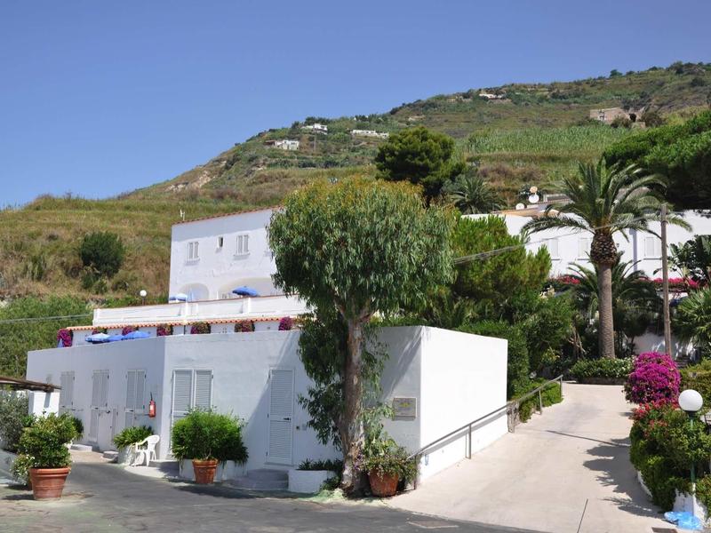 Wit hotelgebouw met terras, omringd door planten en heuvels onder een blauwe lucht.