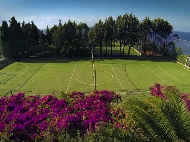 Voetbalveld omgeven door bomen, bloemen en zeezicht onder een heldere hemel.