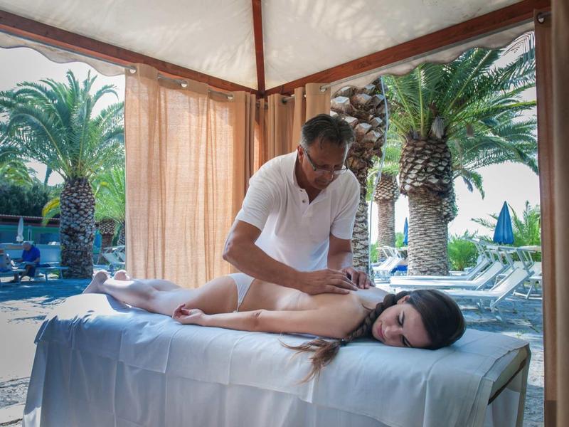Een man masseert een vrouw op een buitentafels onder een overkapping naast palmbomen.