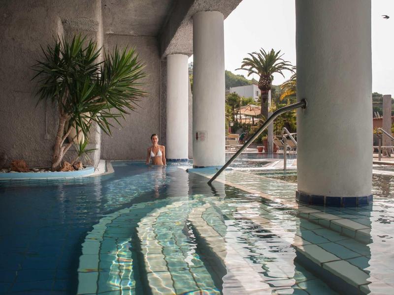 Gebogen zwembadtrappen en zuilen met palm en persoon aan rand in zonnige omgeving.