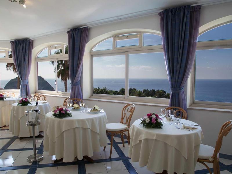 Elegante eetkamer met grote ramen met uitzicht op de zee, tafels met witte tafelkleden en bloemen.