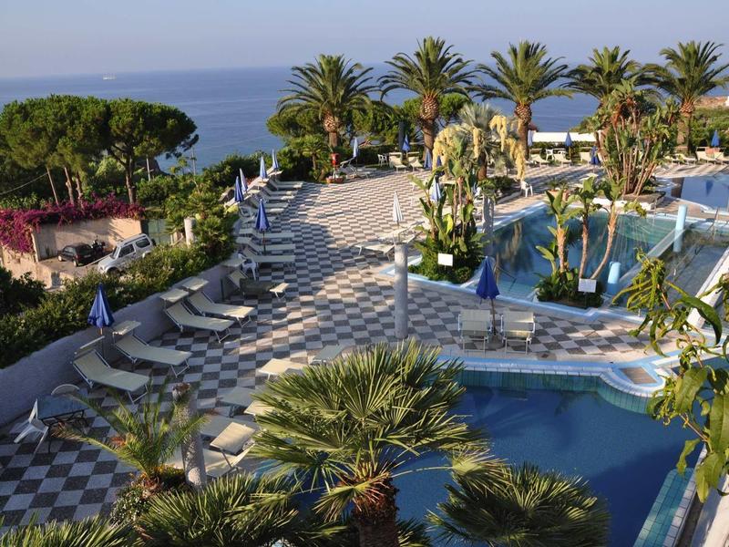 Hotelzwembad met ligstoelen, palmbomen en zeezicht bij heldere lucht.