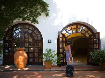 Žena s kufrem vstupuje do budovy s obloukovými dřevěnými dveřmi a kvetoucími rostlinami.