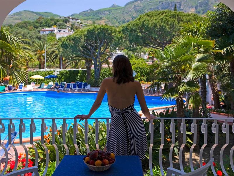 Žena se dívá z balkonu na hotelový bazén s palmami a horami v pozadí.