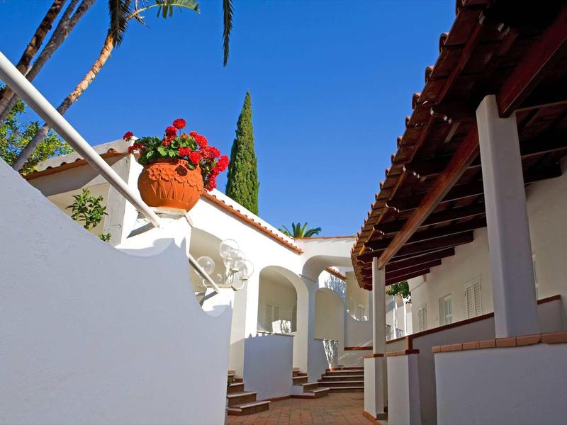 Světlé nádvoří s bílými zdmi, červenými taškami a květinami v hliněném květináči.