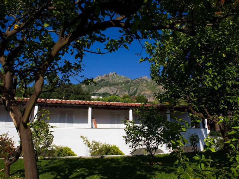 Bílá budova s červenou střechou obklopená stromy a zeleným trávníkem pod jasně modrou oblohou.