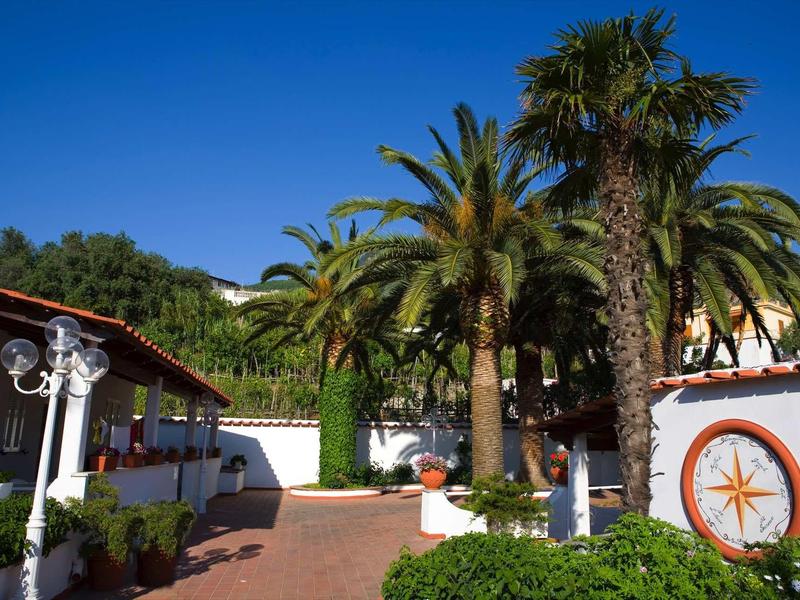 Dvorek s palmami, modrou oblohou a budovou s bílou fasádou a oranžovou výzdobou.