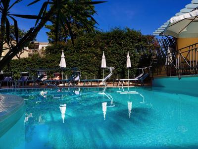 Klares Schwimmbecken mit Sonnenliegen und Sonnenschirmen, umgeben von Palmen und Gebäuden.