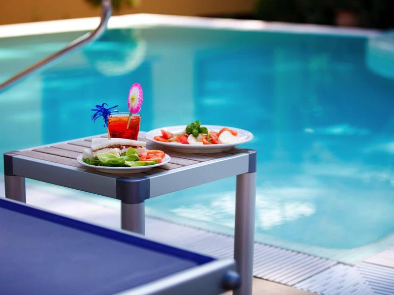 Kleiner Tisch am Pool mit zwei Tellern Essen und einem roten Drink mit Strohhalm und Schirmchen.