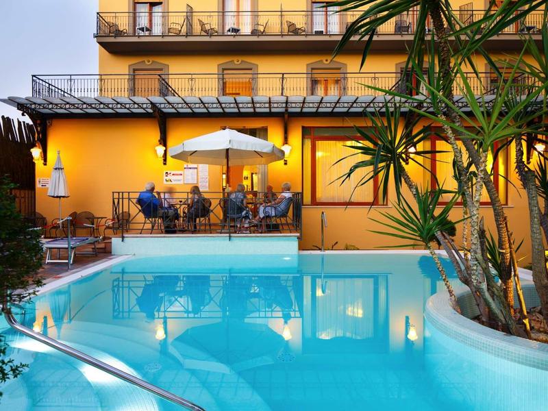 Gelbes Hotel mit Balkon und Außenpool, umgeben von Pflanzen und Sitzbereich mit Sonnenschirm.