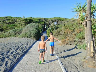 Zwei Jungen laufen auf einem gepflasterten Weg durch eine sandige, bewachsene Landschaft bei klarem Himmel.